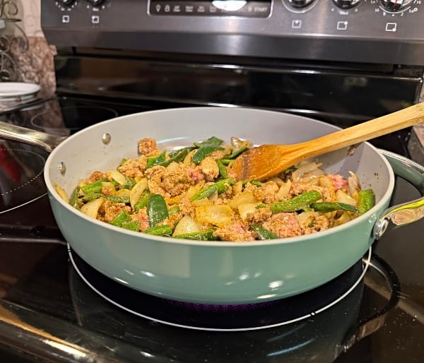 Caraway nonstick pan cooking meal on electric glass top stove with wooden utensil resting in the pan