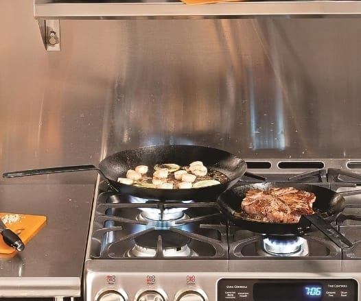 Image-of-two-Lodge-carbon-steel-pans-being-used-to-cook-steak-and-onions-on-gas-stovetop