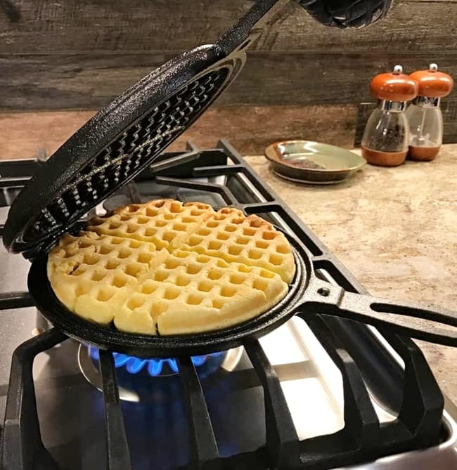 Lehmans-Cast-Iron-waffle-maker-without-nonstick-coating over a gas stove burner with lid half open showing a waffle that's almost finished cooking