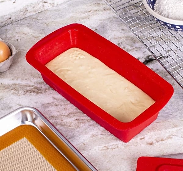 Mrs Anderson's silicone loaf pan filled with batter sitting on a marble countertop with a stainless steel baking sheet in the foreground and cooling rack in the background