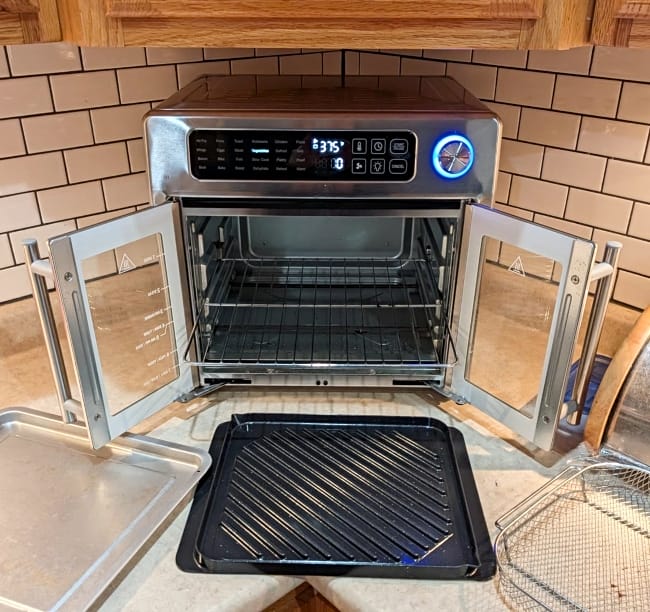 Image of Emeril Lagasse non toxic air fryer oven sitting on countertop with doors open and accessories displayed next to it