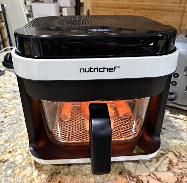 Image of Nutrichef glass and stainless steel air fryer sitting on countertop with egg rolls in the basket cooking under a red interior light
